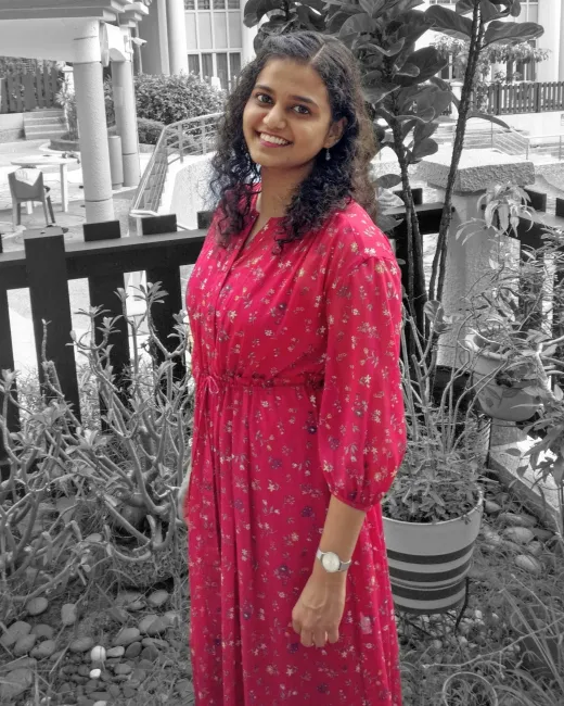 Kavya smiling, in a pink floral dress, among plants and garden chairs in black and white